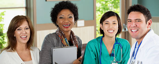 Group of four health professionals smiling, including two women in business attire, a woman nurse with a stethoscope and scrubs, and a man in a business shirt wearing a stethoscope. 