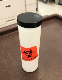 "A white, cylindrical, hard-sided plastic container with a flat, black screw-on lid is placed on a table.  An orange "Biohazard" sticker is affixed to the side of the container."