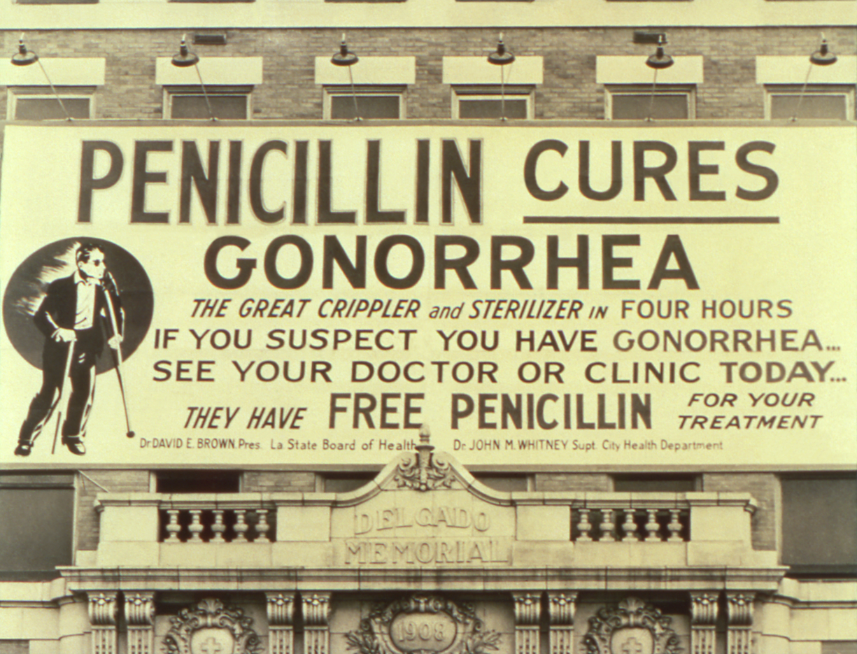 "An historic image of a large billboard hung over the entrance to Charity hospital in New Orleans, Louisiana.  Billboard depicts a World War 2 poster announcing that, “PENICILLIN CURES GONORRHEA, THE GREAT CRIPPLER and STERILIZER in FOUR HOURS; IF YOU SUSPECT YOU HAVE GONORRHEA… SEE YOUR DOCTOR OF CLINIC TODAY… THEY HAVE FREE PENICILLIN FOR YOUR TREATMENT”. The poster also identifies  the parties responsible for its content as, Dr. David E. Brown, Pres. La. State Board of Health, and Dr. John M. Whitney, Supt., City Health Department"