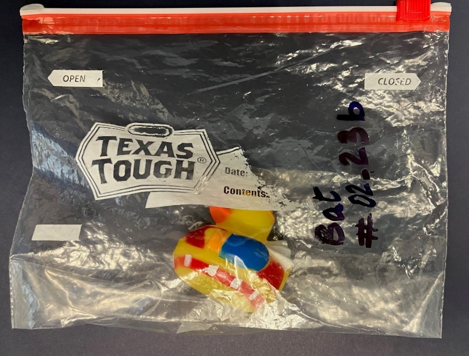 "An overhead image of a closed, transparent zip-seal bag with a toy yellow rubber ducky inside it .  A specimen identification code is written in large dark letter on the zip-seal bag. The identification code reads: Bat #02_23b"