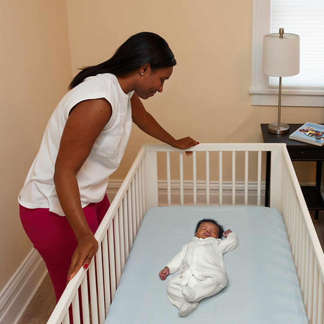 A woman supervising an infant in a crib, with the infant laying on their back to sleep.