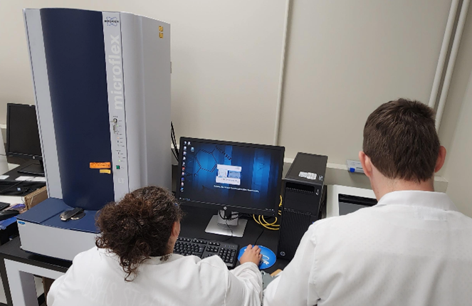 Two people in lab coats working on a computer beside an instrument (MALDI-ToF analyzer)