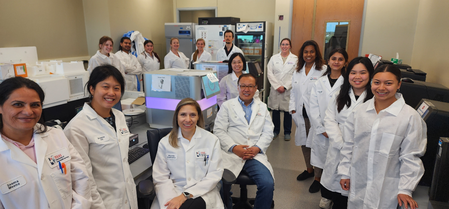 A group of 16 people in lab coats smiling and standing next to sequencing instruments. 