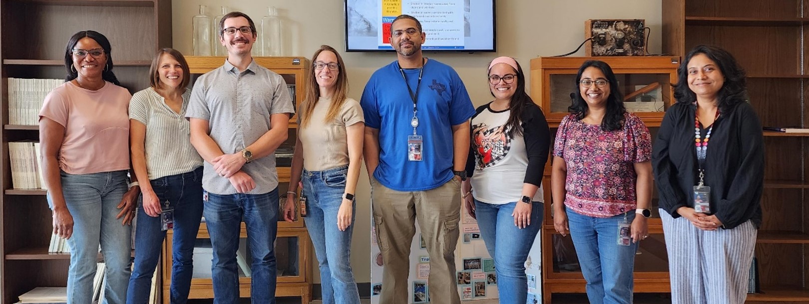 A group of eight people standing together smiling in front of shelves and cabinets.
