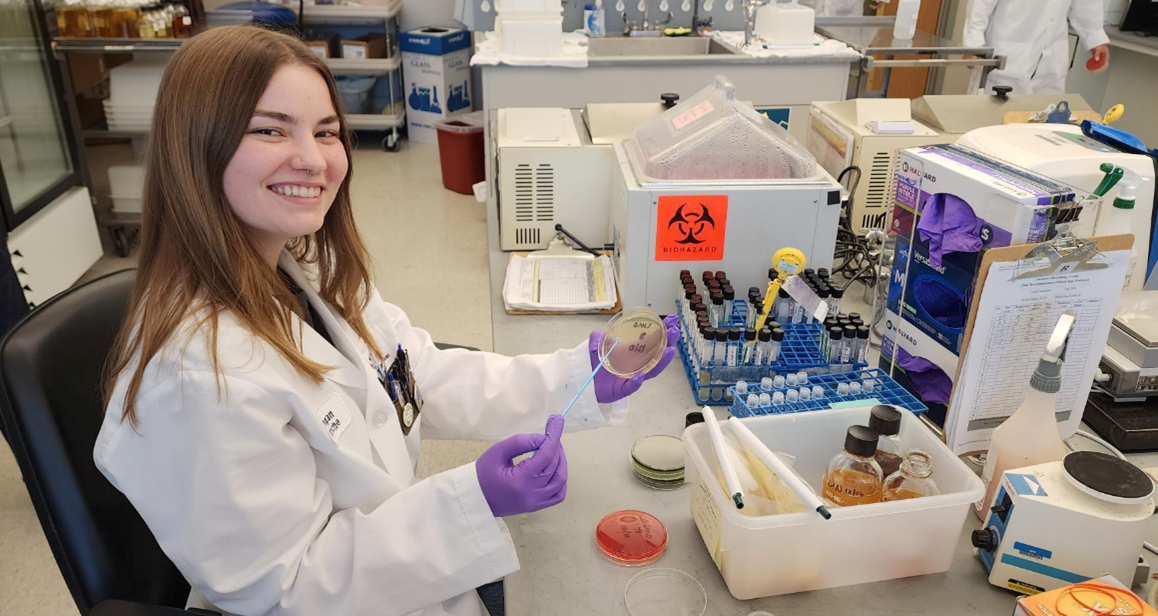 A person in a lab coat holding a petri dish in one hand and streaking it with an inoculation wand.