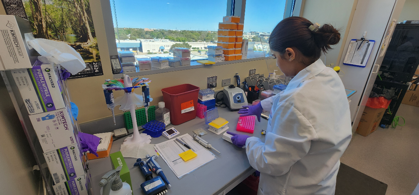 A person in a lab coat working at a laboratory bench next to several micro pippettes.