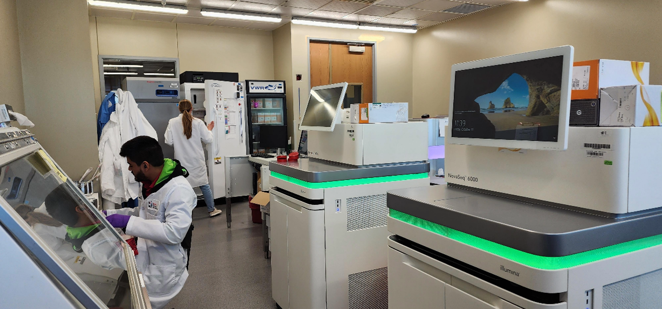 A room with several sequencers and fridges. Two people in lab coats in the background are working on the instruments.  