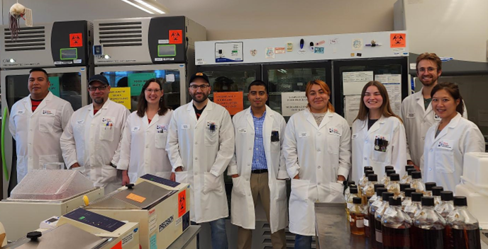 A group of nine smiling people in lab coats standing in a laboratory