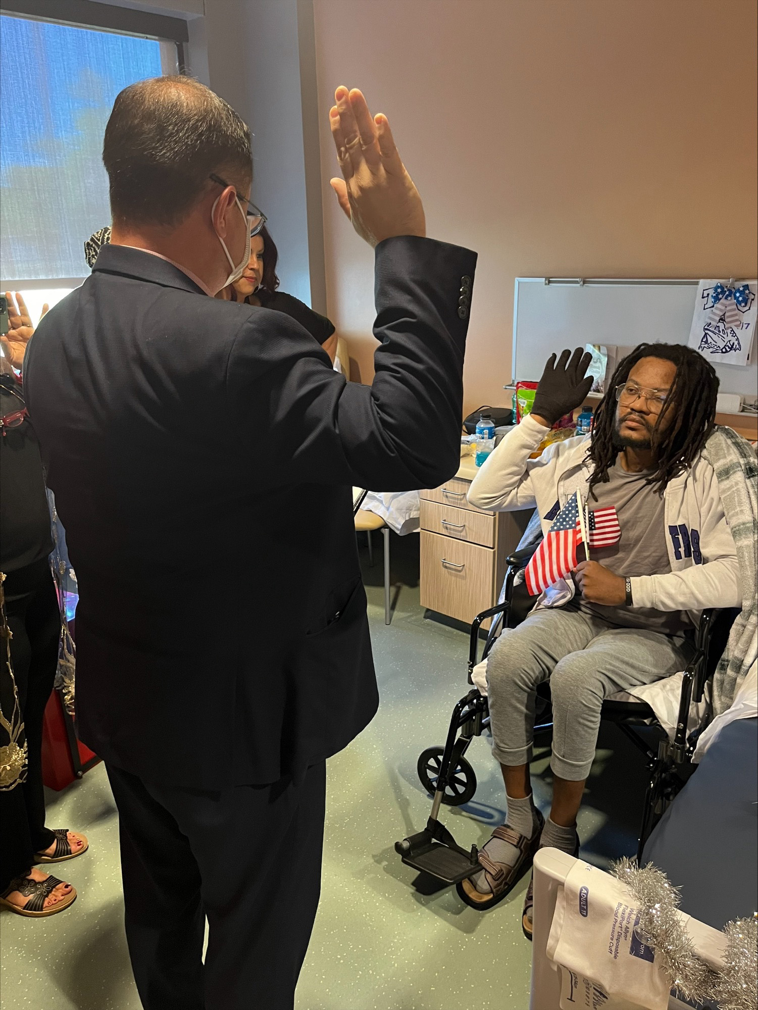 A man in a suit leads another man in a wheelchair holding an American flag in an oath-taking ceremony for US citizenship.