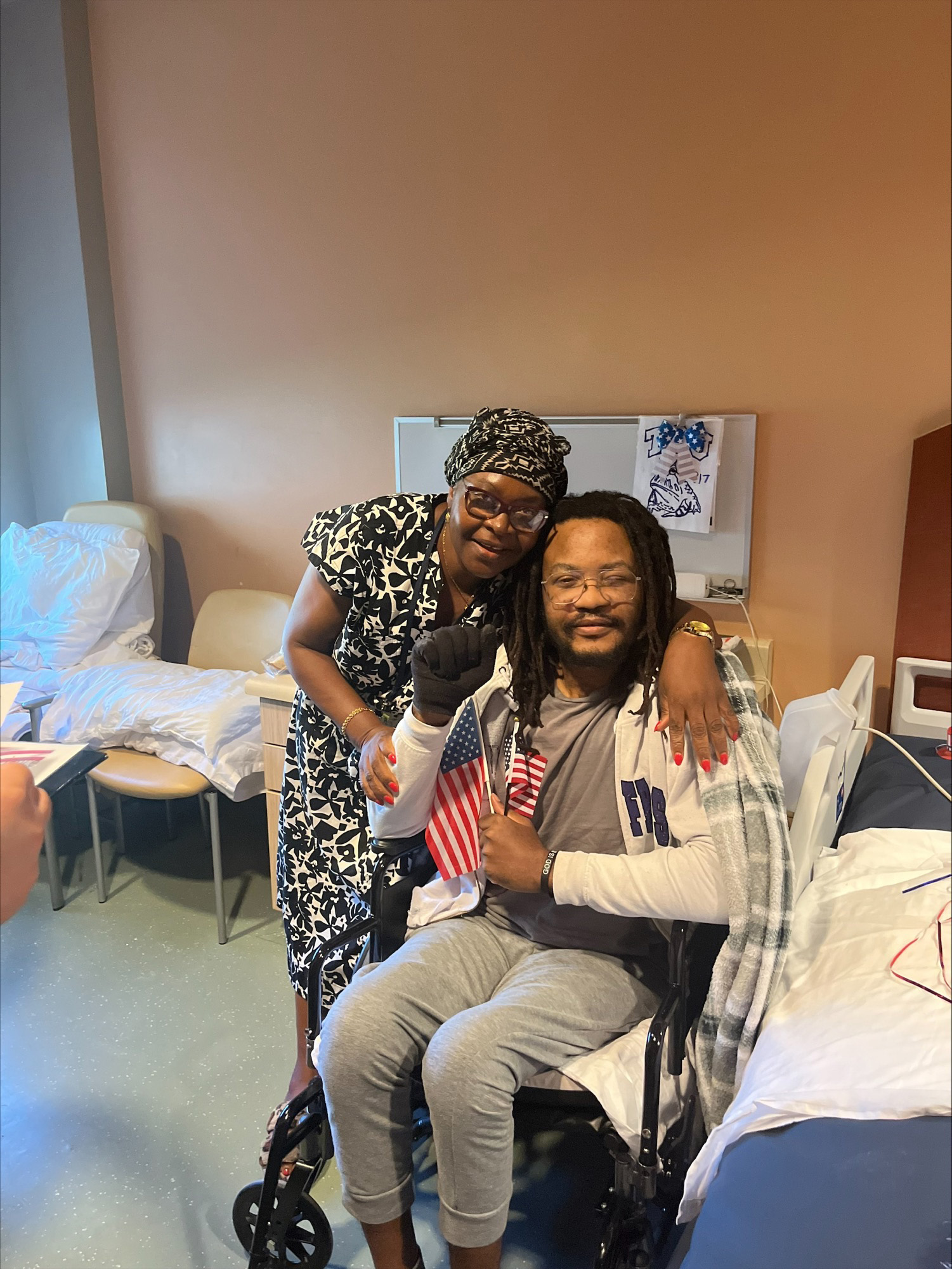 A man in a wheelchair holding an American flag with a woman smiling next to him in a hospital room.