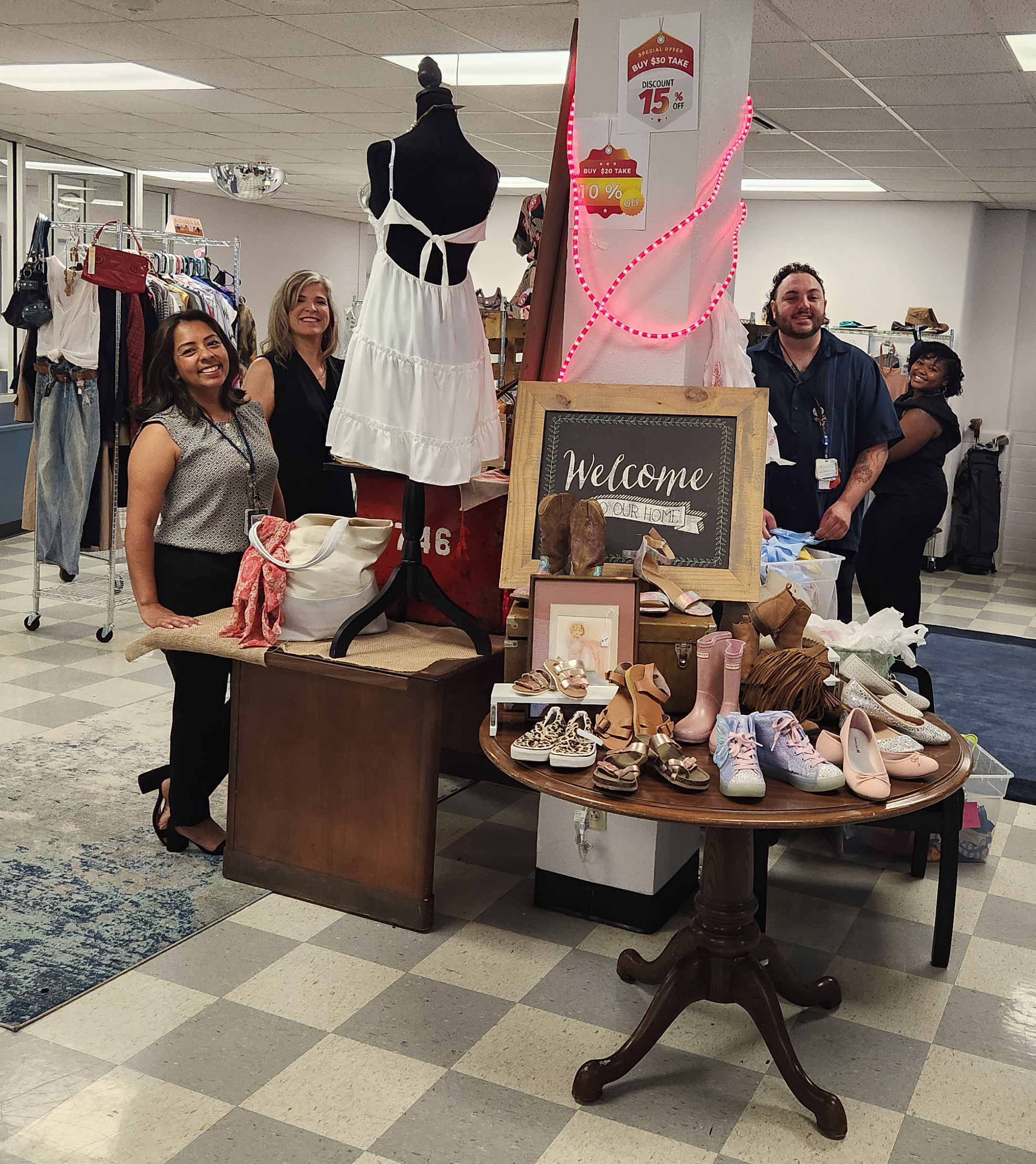 People posing in a thrift store with a display of children's shoes, a mannequin, and a welcome sign.
