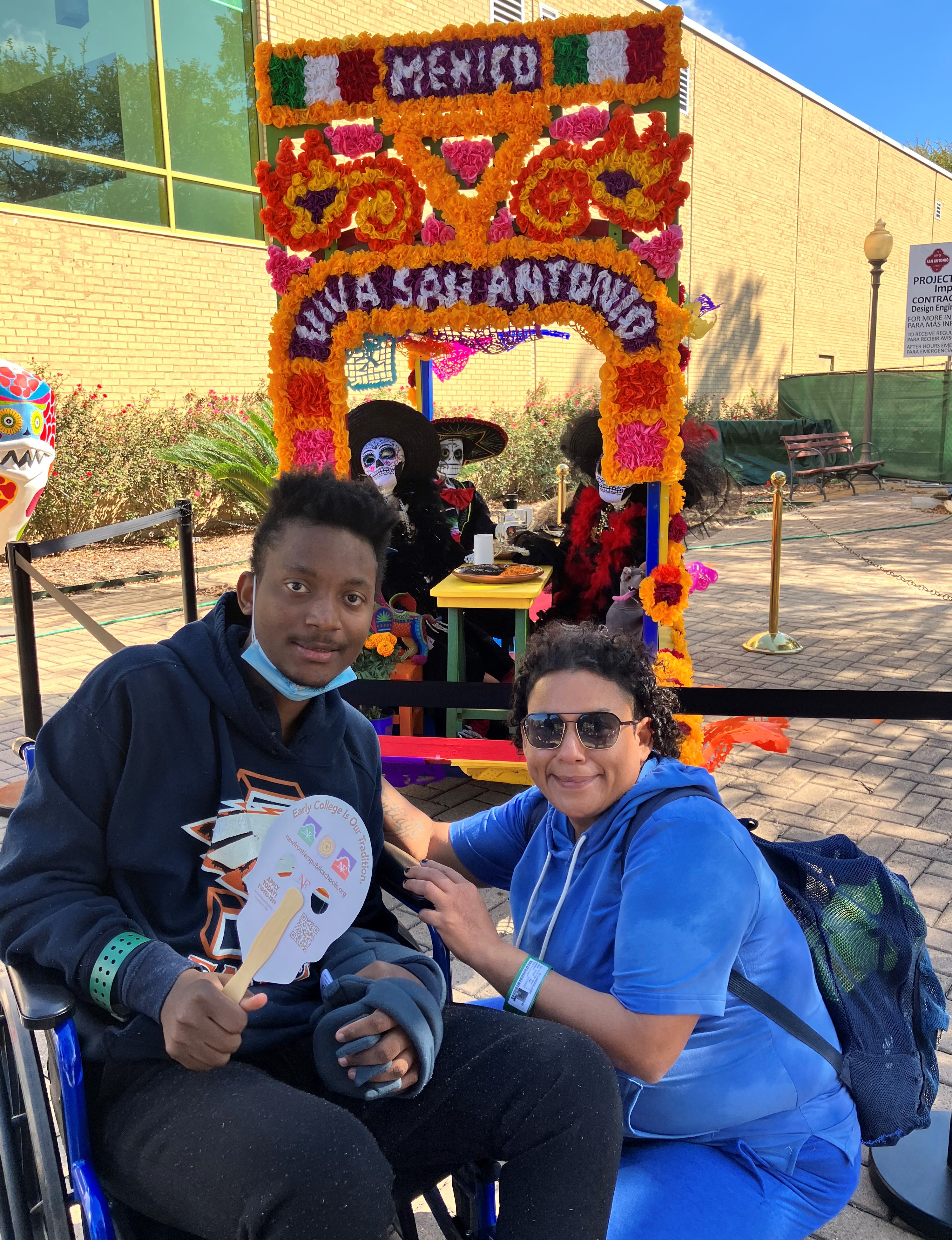 Two people in front of a colorful Día de los Muertos display with floral decorations and Catrina figures.