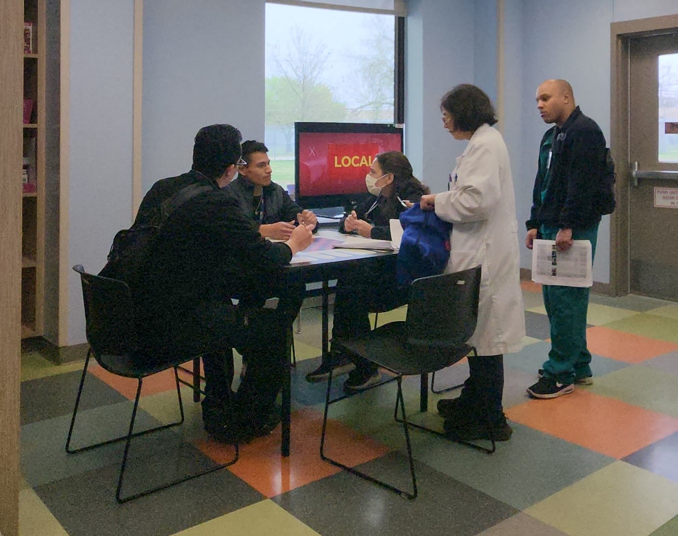 A group of health professionals sitting and standing around a table in a room with a colorful tiled floor and a television.