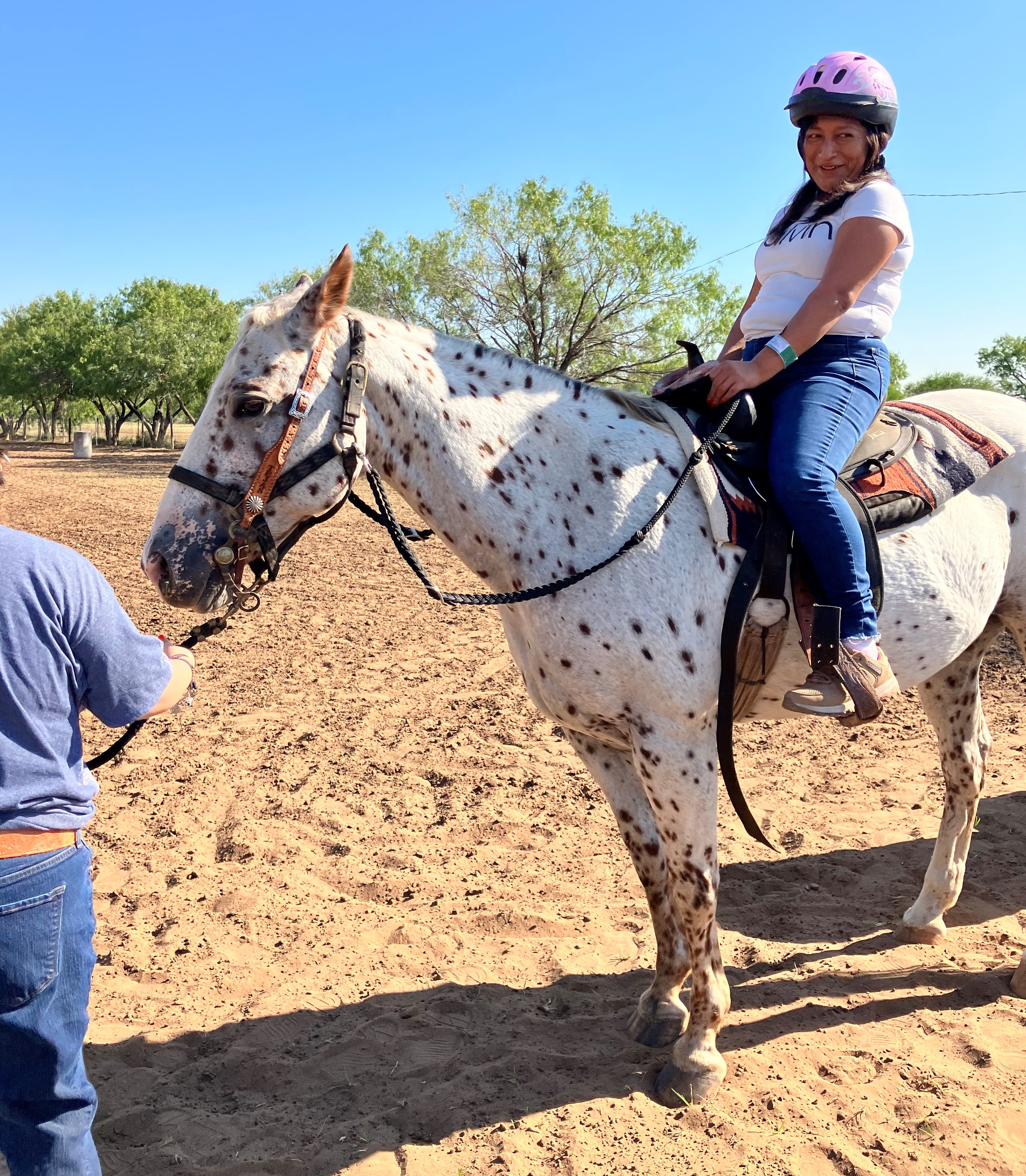 Person riding a white horse with dark spots, wearing a pink helmet. Another person holds the reins on the ground.