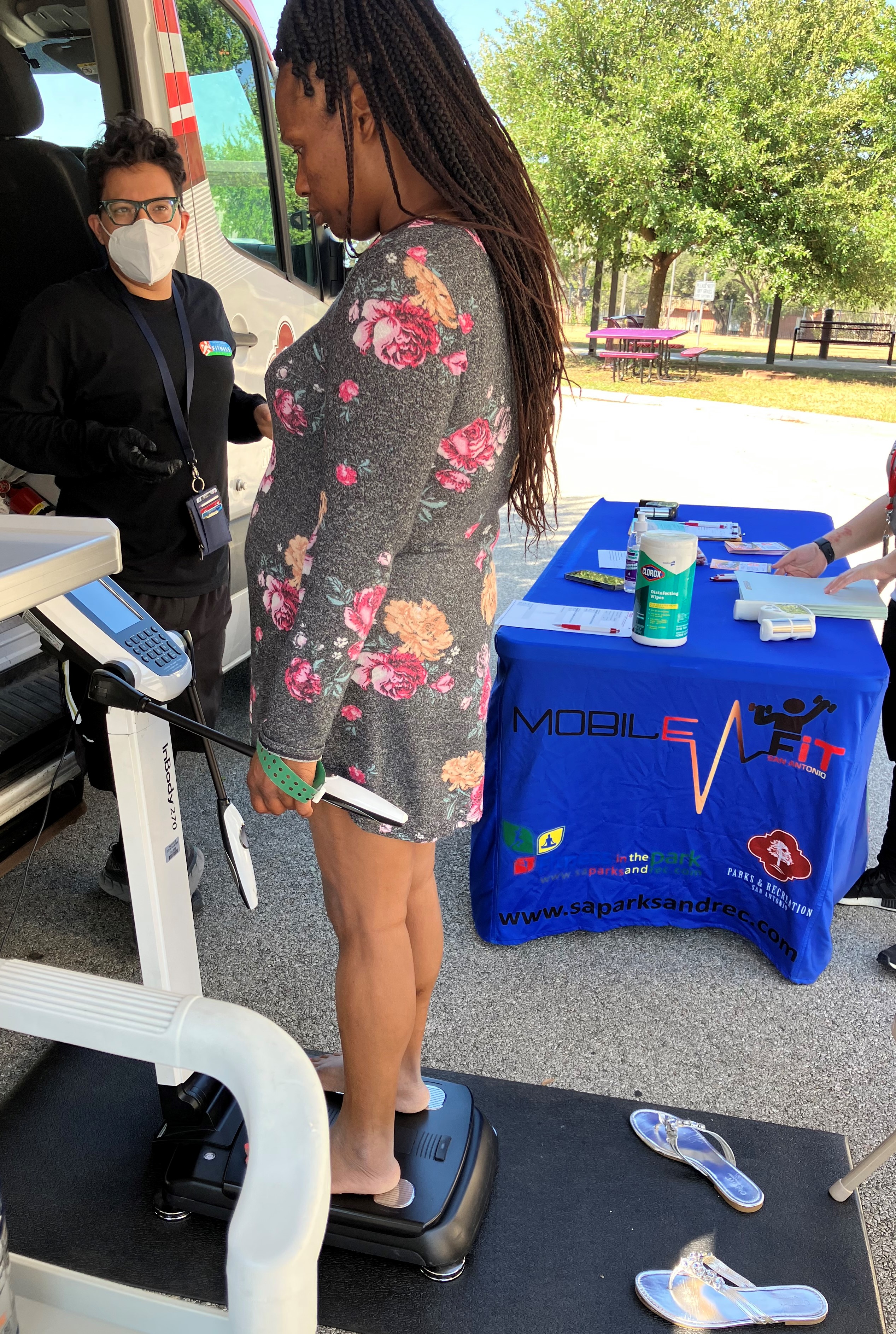 Una persona se encuentra de pie sobre una báscula de composición corporal al aire libre, mientras otra persona observa a su lado, junto a una furgoneta de servicios de salud.