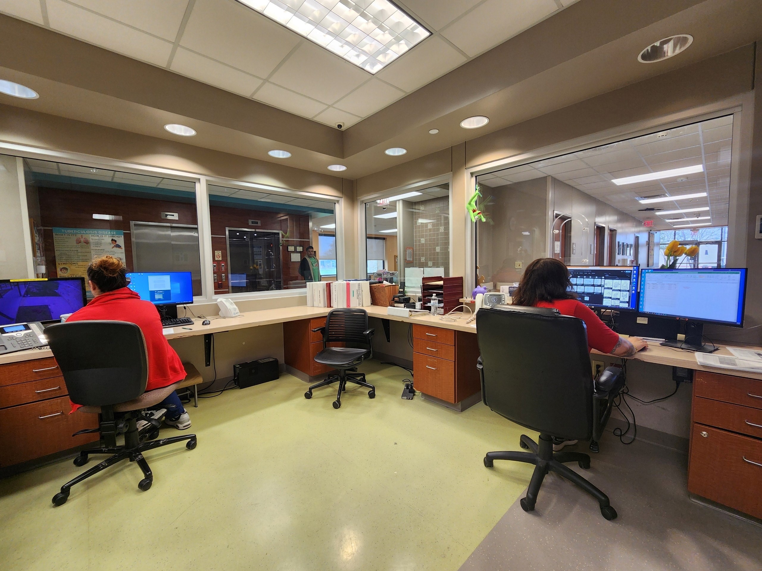 Nurse station with two people working at computer workstations, separated by a large interior window.