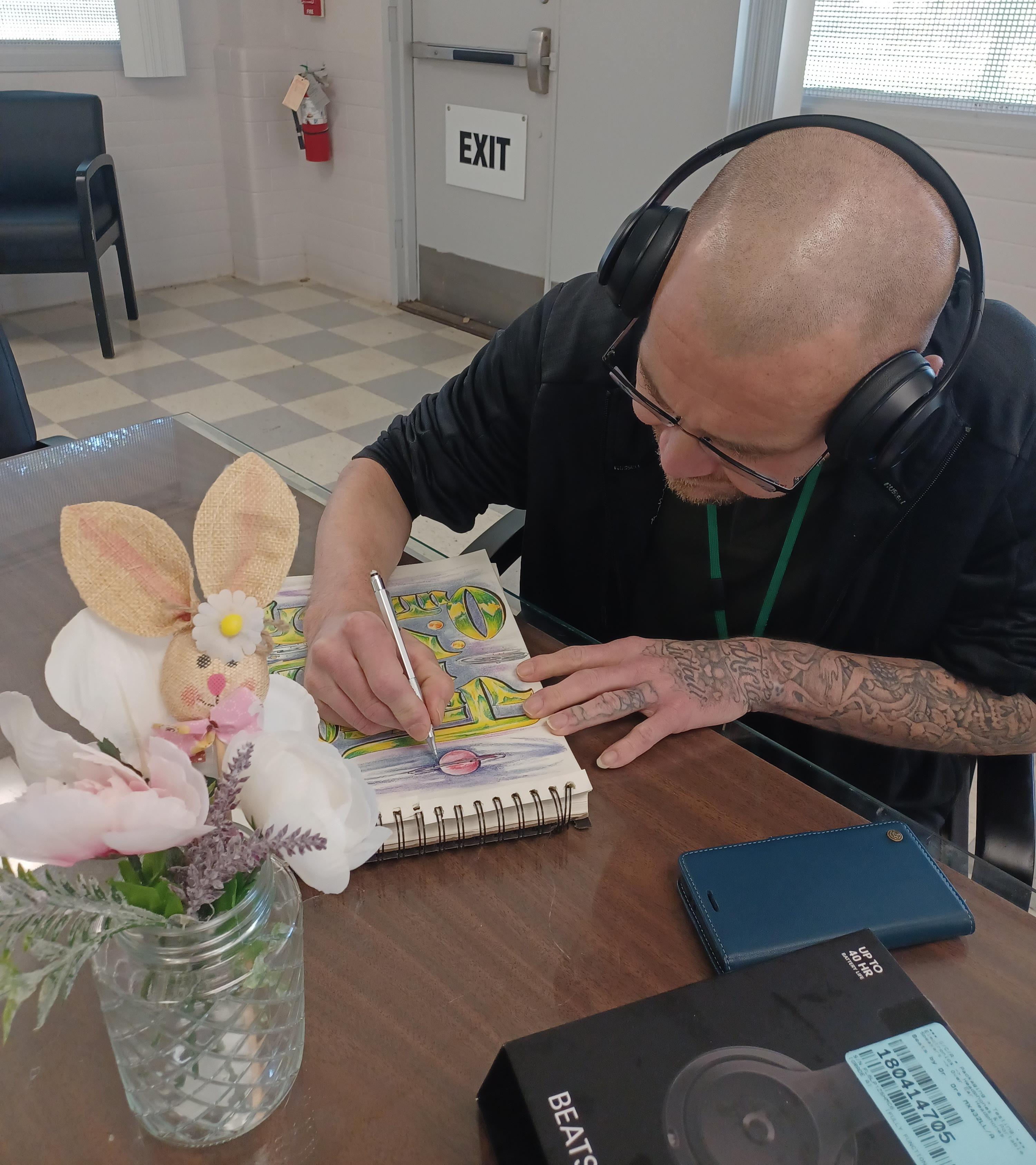 A person wearing headphones drawing in a sketchbook at a table with a floral decoration and a Beats box nearby.