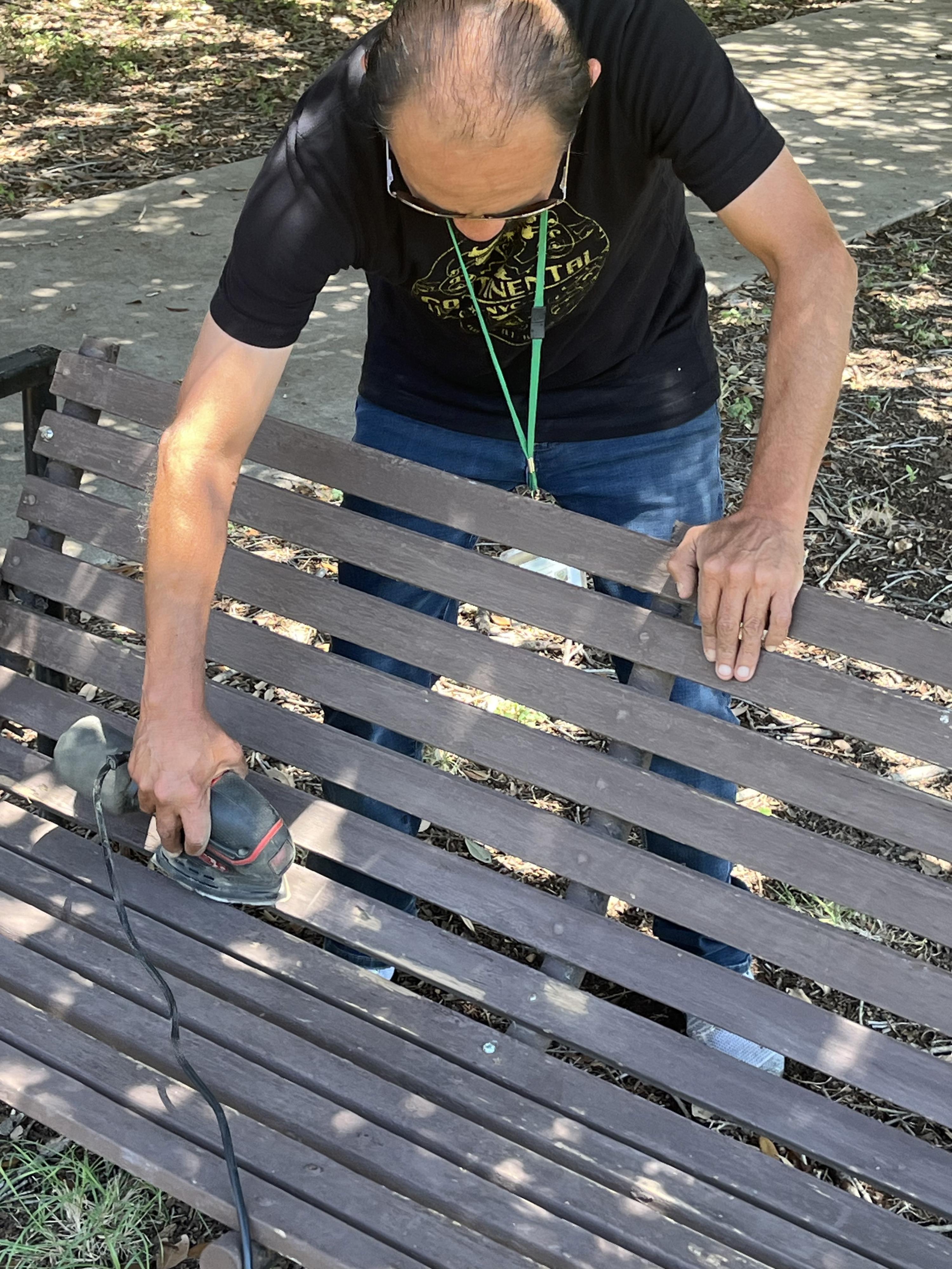 A man sands a wooden bench with a tool