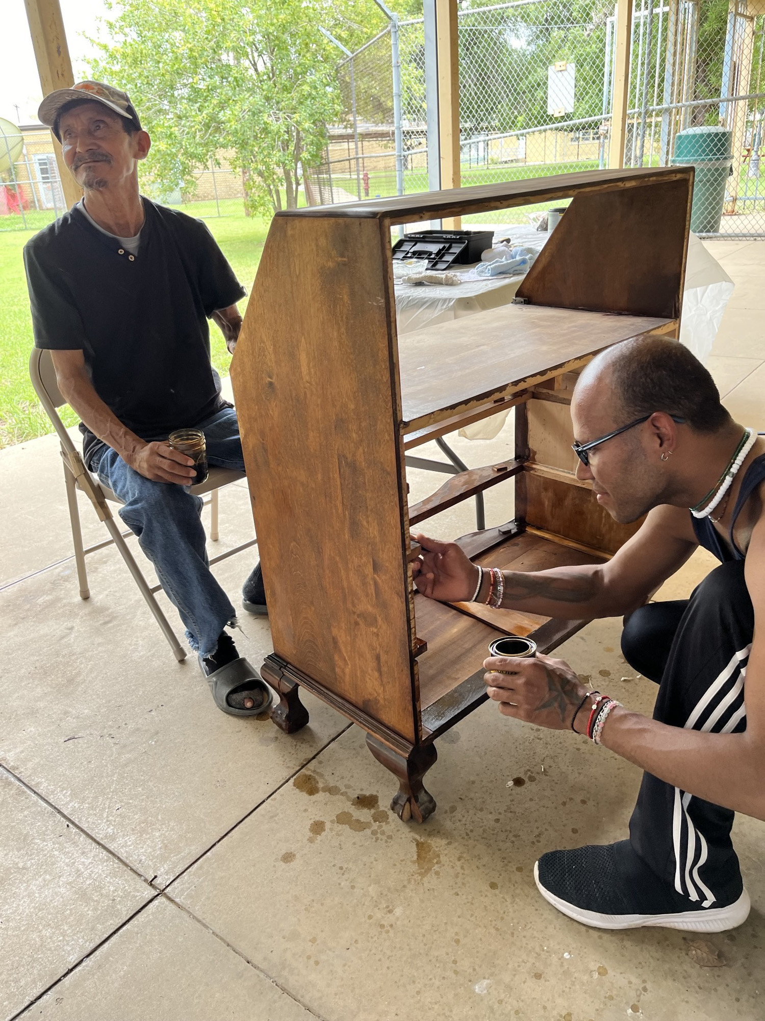 Two people varnishing a wooden cabinet outdoors on a patio.