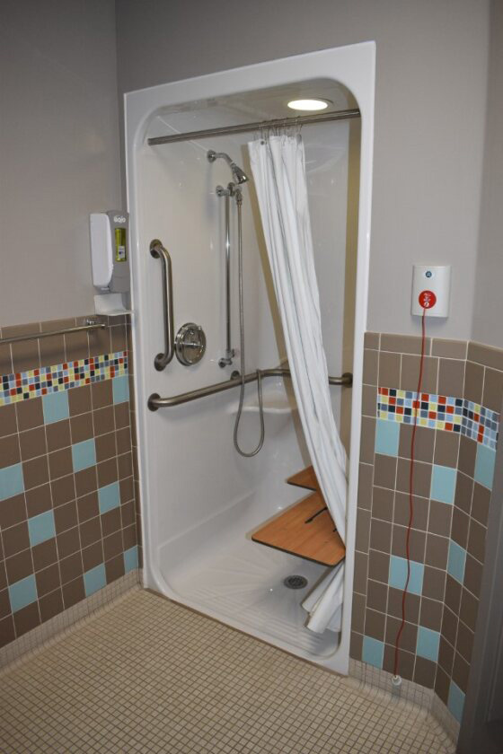 Accessible shower unit with metallic grab bars, a wooden seat, and a handheld showerhead, surrounded by beige and mosaic tile walls.