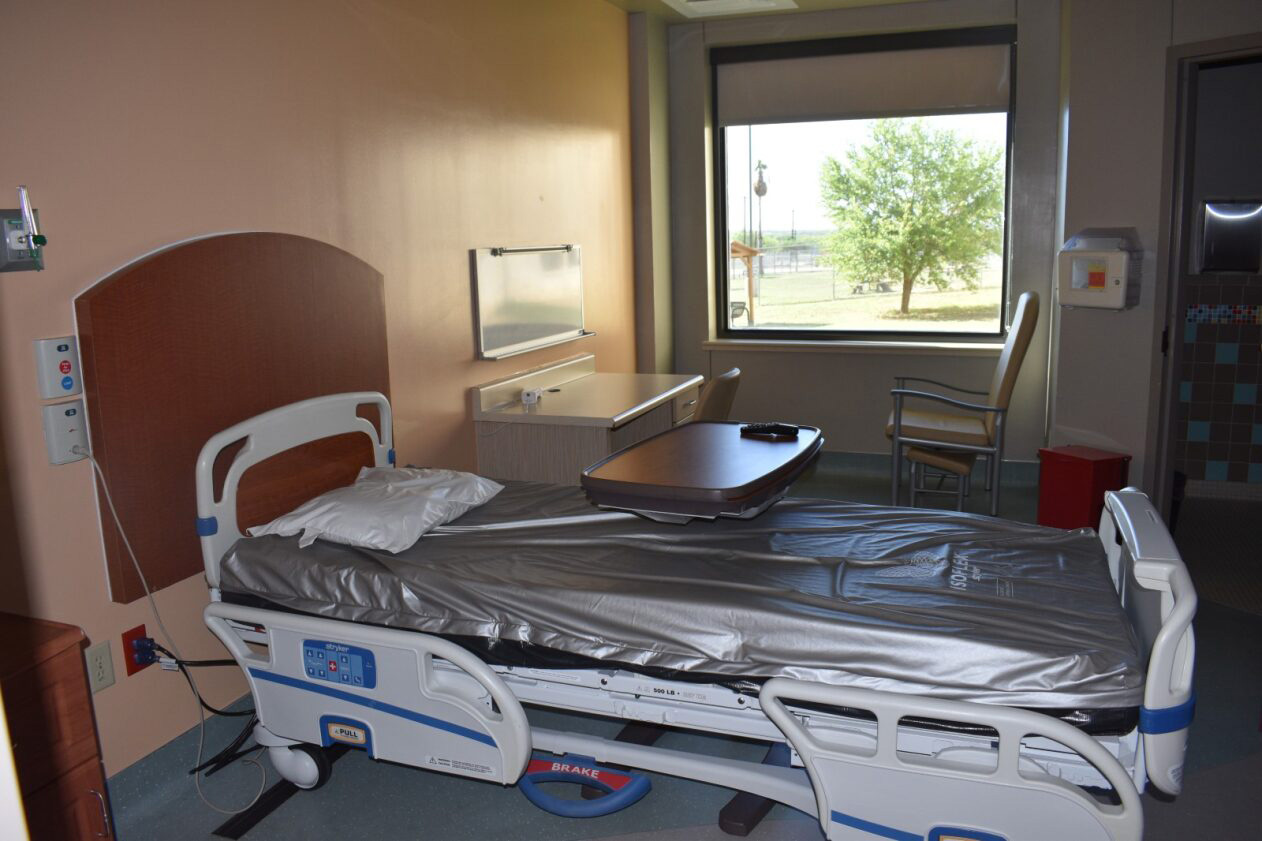 Hospital room with a bed, desk, chair, armchair, and a large window showing a tree outside.