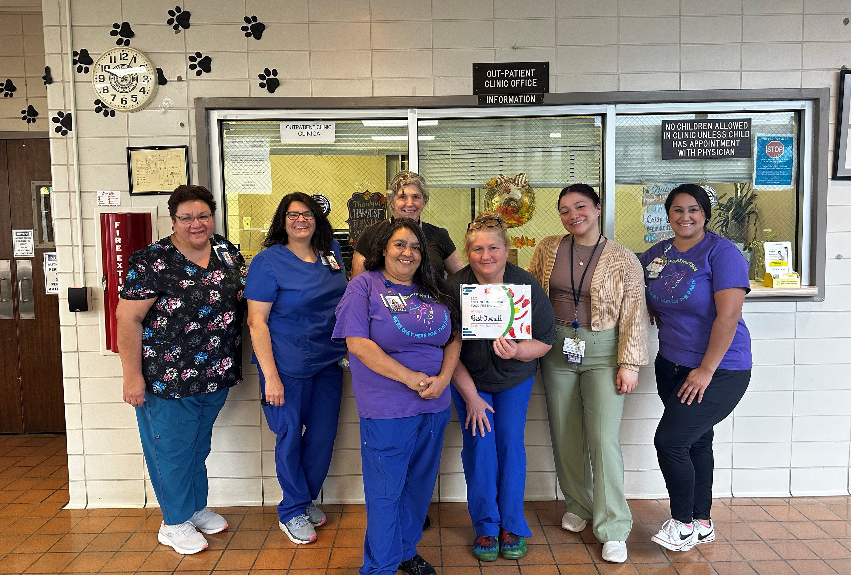 A group of seven people poses in front of a clinic office window, smiling, with a certificate held by one person.