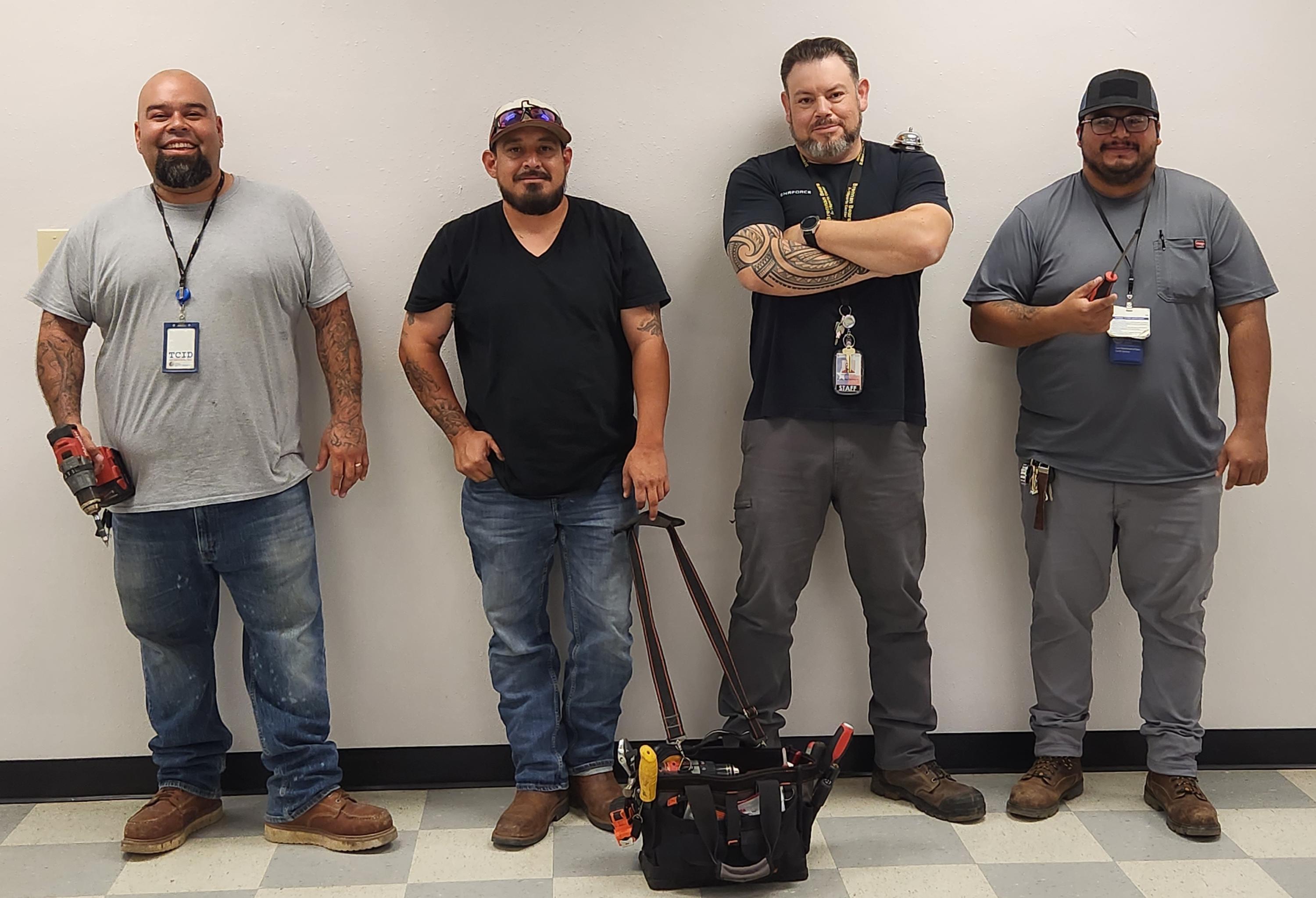 Four men in work attire standing against a wall, with tools in hand and a tool bag on the floor.