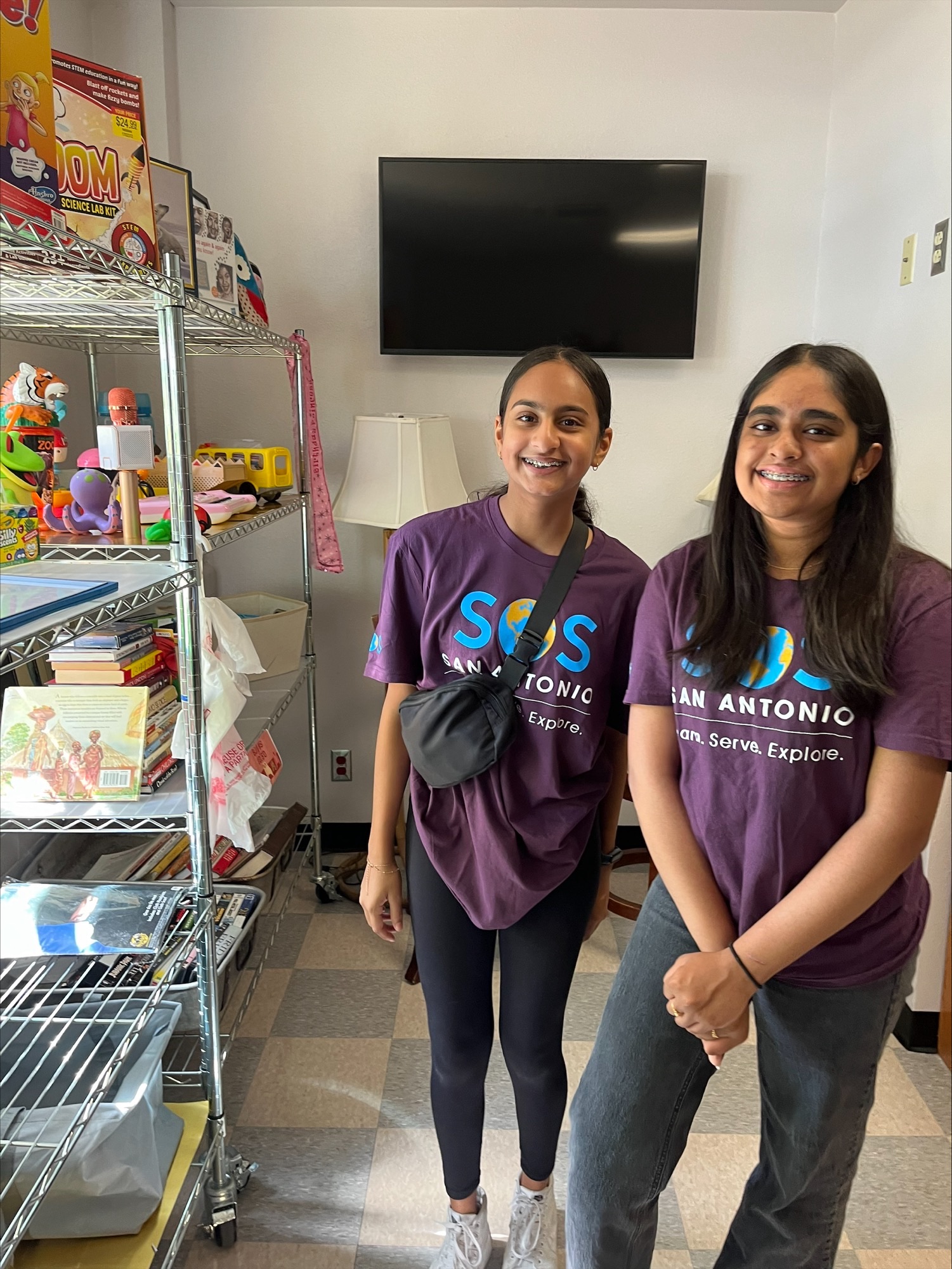 Two people in purple shirts stand in a room with shelves of toys and a wall-mounted TV.